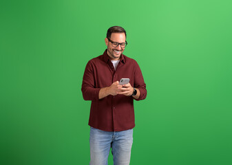Handsome young man in eyeglasses smiling and texting online over smart phone on green background