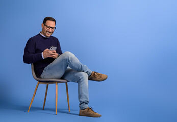 Handsome young man smiling and using social media over smart phone on chair against blue background