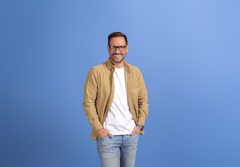 Portrait of handsome young man in eyeglasses and with hands in pockets standing on blue background