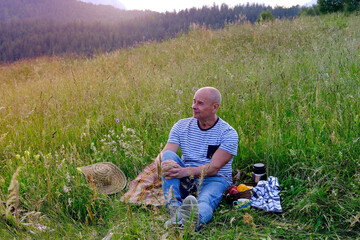 mature man, senior sits on gentle slope of mountain, next to picnic basket with fruit, thermos,...