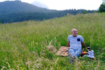 mature man, senior sits on gentle slope of mountain, next to picnic basket with fruit, thermos,...