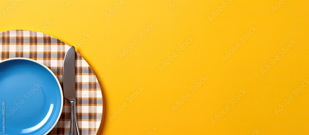 Poster copy space image of a yellow background with an empty plate cutlery a blue checkered napkin and a mu