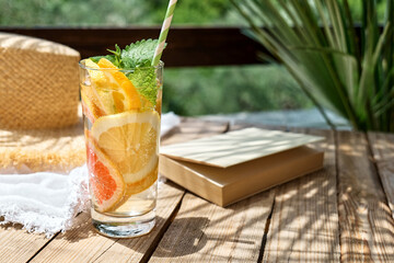 Summertime mood. Citrus refresh drink, book and hat on white beach blanket in tropical garden.