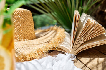 Summertime mood. Citrus refresh drink, book and hat on white beach blanket in tropical garden.