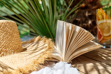 Summertime mood. Citrus refresh drink, book and hat on white beach blanket in tropical garden.
