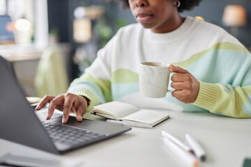 Minimal closeup of Black young woman typing at laptop keyboard in office and holding coffee mug...