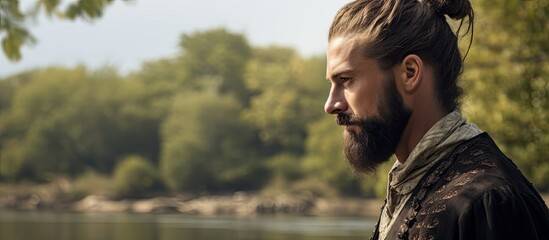 A bearded man with a stylish hair bun and make up is seen turned to the side looking away while speaking with a picturesque background of a tree and a river. Copyspace image