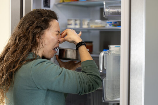 Woman holding her nose while taking spoiled food out of the refrigerator. Bad smell in the refrigerator due to spoiled food. Woman holding her nose due to unpleasant bad smell of spoiled food.