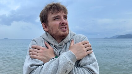 Young man shivering and trembling from cold rainy weather at summer vacation on beach, sea