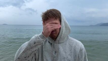Young man shivering and trembling from cold rainy weather at summer vacation on beach, sea