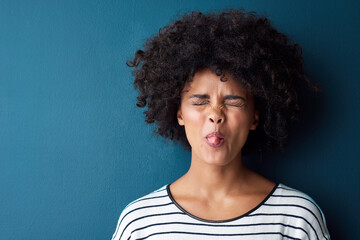 Portrait, emoji and tongue with facial expression in studio for fun, relax and playful attitude on blue background. Lady, comic and goofy black woman with a funny face, crazy and personality isolated