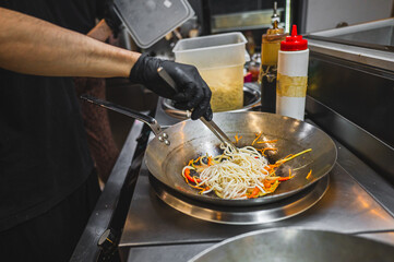 Close-up of a chef’s hands cooking stir-fry noodles with vegetables in a wok on a stove,...