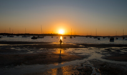 Silhouette of a person walking in the sand against the sea with boats at sunset