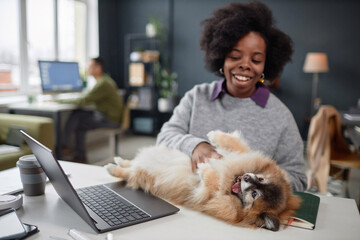 Portrait of smiling African American woman playing with cute dog while working at desk in pet...