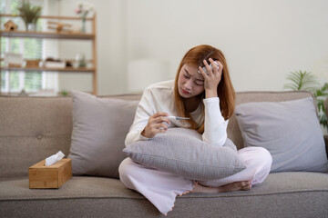 Young Asian woman feeling unwell and checking her temperature with a thermometer while sitting on the sofa at home. Concept of illness, fever, and healthcare