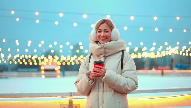 Woman in earmuffs and warm clothes holding takeaway coffee standing outside ice rink. Portrait of beautiful lady smiling at camera against blurred Christmas lights in city during evening.
