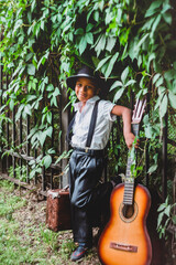 a small guitarist in a hat with a guitar and a suitcase stands leaning against an iron fence covered with ivy