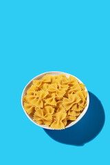 Uncooked bow tie pasta, farfalle in a white bowl on a vibrant blue background, perfect for culinary concepts.