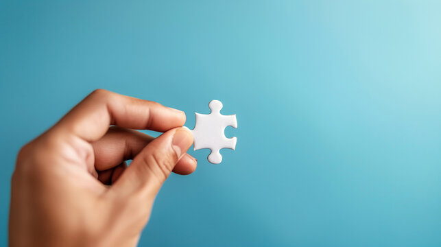 A close-up of a hand holding a single white puzzle piece against a plain blue background, symbolizing problem-solving, teamwork, and the last piece of the puzzle.