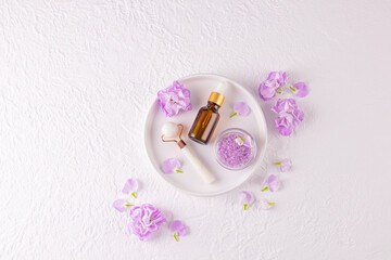 Cosmetic set for spa and skin care, massage. Stone roller, bath salts, massage oil on ceramic white tray. top view. lilac delicate flowers