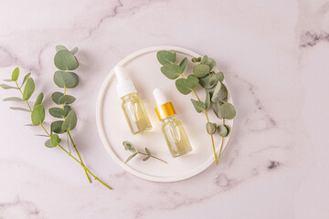Two cosmetic bottles with a dropper with natural eucalyptus oil and a product for the care of problem skin on a white plate with eucalyptus branches.