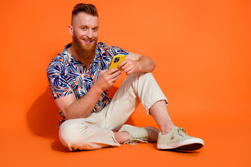 Full size photo of cheerful man wear stylish shirt pants sitting with smartphone in hands chatting...