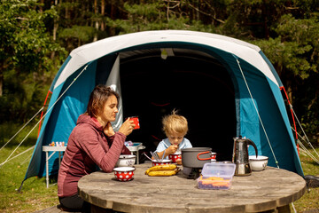 Family, mother and child, having breakfast in front of pitched tent in forest, while wild camping in Norway, summertime