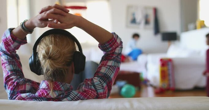 A mother sits in the foreground with noise-canceling headphones, peacefully detached from her children's playful chaos in the background.