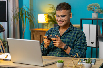 Excited Indian Hindu man playing video game on laptop spending leisure time at home office desk....
