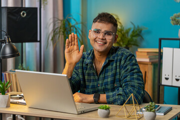 Hello. Indian man smiling friendly at camera and waving hands gesturing hello, hi, greeting or...