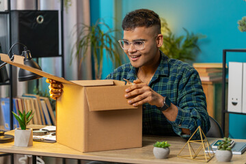 Happy Indian man unpacking delivery parcel. Smiling satisfied guy shopper online shop customer opening cardboard box receiving purchase gift by fast postal shipping at home office desk. Lifestyle