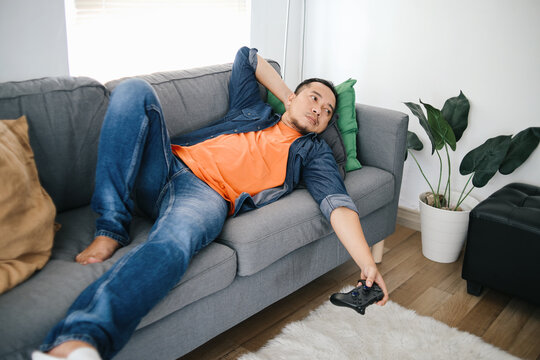 Bored man holding joystick, playing video games while lying on sofa at home