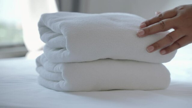 Close-up footage of a woman's hand placing a stack of fresh white towels on the bed. The unidentified woman is laying the folded clean towels on the bed sheet in the room.
