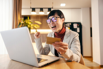 An excited japanese woman in formal wear with credit card in her hand is celebrating while looking at the laptop.
