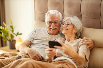 Romantic senior couple is sitting on a bed in a bedroom and taking selfies.