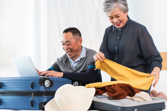 Happy Asian senior couple preparing for journey. Old man using laptop to planning and woman packing suitcases with excitement for trip, smiling, sitting on bed at home, getting ready vacation together