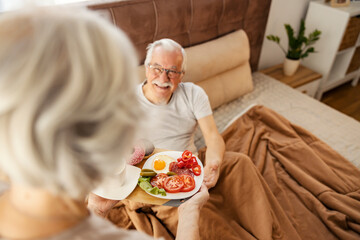 Romantic senior woman is bringing breakfast in bed to her beloved husband.