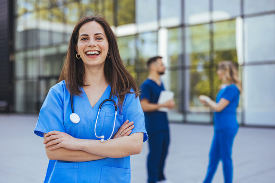 A cheerful Caucasian female nurse, clad in blue medical attire with a stethoscope, stands confidently outside a hospital as colleagues converse in the background.