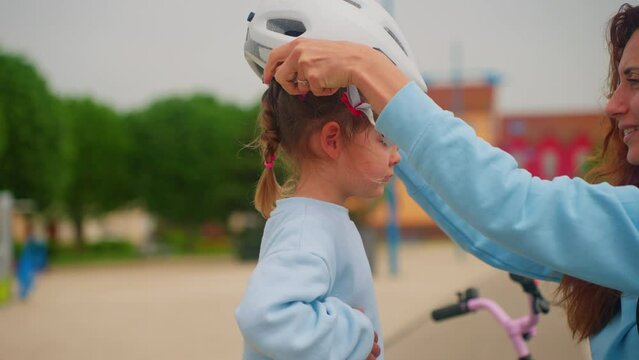 Loving mother put on sport helmet to little cute kid daughter before spin bicycle on street. Little girl wear protective halmet for health care and safety
