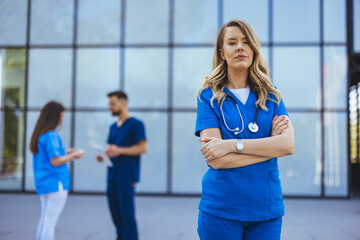 A confident Caucasian female nurse in blue scrubs stands with arms crossed in front of a hospital...