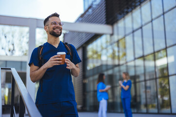 A cheerful male healthcare worker in blue scrubs sips coffee outside a medical building, with diverse colleagues chatting in the background.