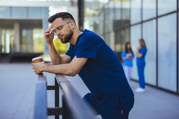 A male nurse in blue scrubs leans wearily on a railing outside a medical facility, holding a coffee...