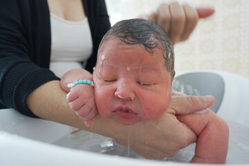 Mother bathing newborn baby in a white tub, baby wearing a name bracelet, eyes closed, water...