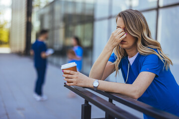 Fototapeta premium A tired female healthcare worker in scrubs holds coffee and rubs her eyes, signaling exhaustion, as colleagues converse in the background outside a medical facility.
