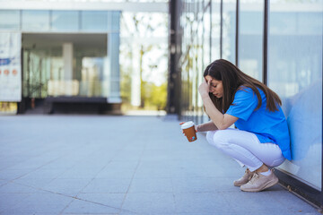 A female healthcare professional sits outside a clinic, appearing exhausted, as she holds a coffee...