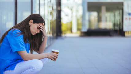 A young female healthcare professional sits outside a medical facility, looking worn out with a...