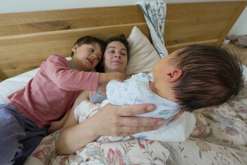 Mother lying in bed with her newborn baby and older child, interacting lovingly, highlighting the...