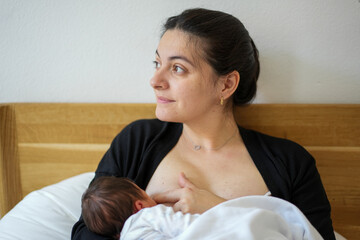 Mother breastfeeding her newborn baby while sitting on a bed. The baby is cradled in her arms,...