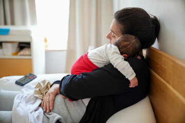 Side view of a mother holding her newborn baby close to her chest while sitting on a bed. The baby...