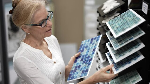 Happy mature woman touching glass wall tiles while shopping through samples for home redecorating renovation project, in a hardware store.